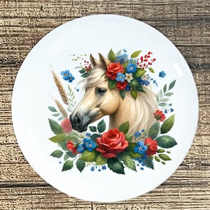 May include: White ceramic plate featuring a horse's head adorned with a floral crown of red roses, blue forget-me-nots, and green leaves. The horse has a cream-colored coat and a flowing mane. The plate has a glossy finish.