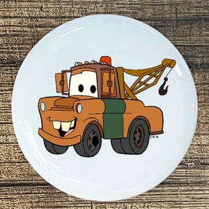 May include: A cartoon illustration of Mater, the tow truck from the Disney Pixar movie Cars, on a white round button. Mater is brown with a green stripe and a toothy grin. He has a large hook on the back of his tow truck.