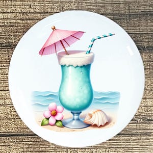 May include: A white ceramic plate featuring a tropical cocktail with a pink umbrella and a striped straw. A pink flower and seashell are on the sand, with ocean waves in the background. The cocktail is light blue.