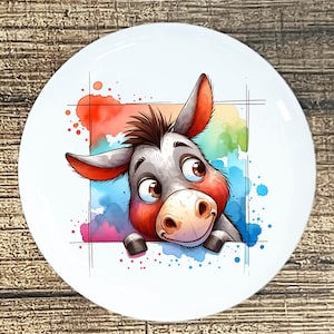 May include: White ceramic plate featuring a cartoon donkey with a watercolor background. The donkey has gray fur, red ears, and a brown mane. The background has splashes of red, blue, and green.