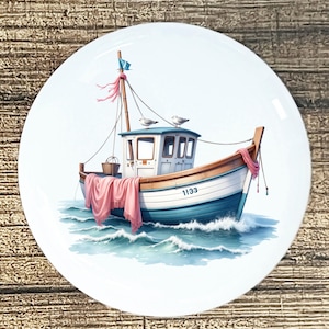 May include: A white ceramic plate featuring a watercolor illustration of a fishing boat at sea. The boat is blue and white with a pink cloth draped over the side. Two seagulls perch on the roof. The boat has the number 1133 on the side.