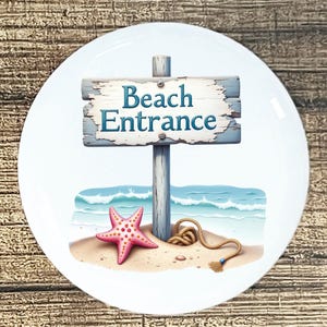 May include: A white ceramic plate featuring a beach scene. The image includes a sign that reads "Beach Entrance" in blue text, a pink starfish, and a rope on a sandy beach with ocean waves in the background.