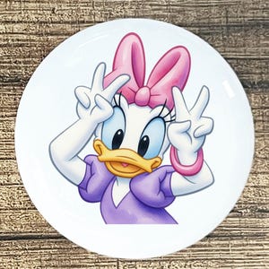 May include: A white ceramic plate featuring a cartoon image of Daisy Duck. Daisy is depicted with a pink bow, purple dress, and is making peace signs with her hands. The plate has a glossy finish and is set against a wooden background.