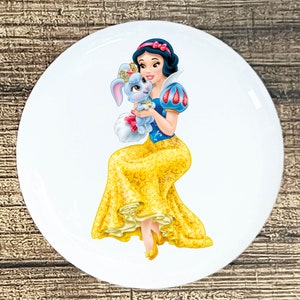May include: Snow White, a Disney princess, is wearing a yellow and gold dress with a blue bodice and a red bow in her hair. She is holding a small, white rabbit with a blue fur coat and a crown.