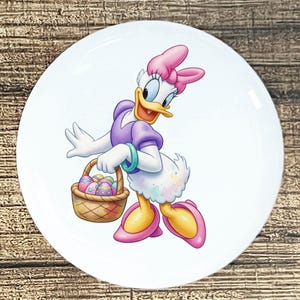 May include: White ceramic plate featuring Daisy Duck from Disney, holding a basket of Easter eggs. Daisy is depicted in a purple dress, pink bow, and shoes, with a turquoise bracelet. The plate has a glossy finish and is set against a wooden background.