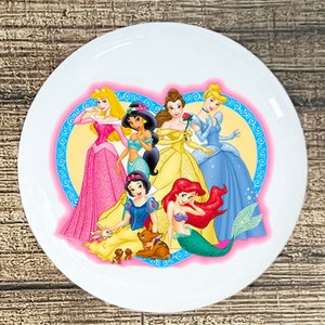 May include: A white ceramic plate with a pink heart-shaped design featuring seven Disney princesses: Snow White, Cinderella, Ariel, Belle, Jasmine, Aurora, and Mulan. The princesses are surrounded by glitter and floral details.