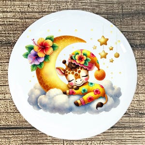 May include: A white ceramic plate featuring a whimsical illustration of a sleeping giraffe wearing a nightcap and pajamas, resting on a cloud next to a crescent moon. The design includes floral accents and hanging gold stars.