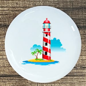 May include: A white ceramic round plate with a red and white striped lighthouse on a small island with a palm tree. The lighthouse has a white roof and a blue sky behind it.