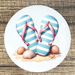 May include: A white ceramic plate featuring a beach scene. Two blue and white striped flip-flops with pink straps are set on a sandy beach with seashells and a starfish. The plate has a glossy finish.