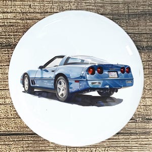 May include: A blue sports car illustration on a white ceramic plate. The car is a classic model, viewed from the rear, with red taillights and a license plate. The plate has a glossy finish and is set against a wooden background.