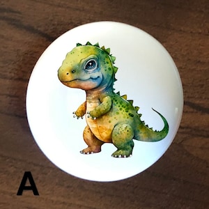 May include: A white ceramic knob with a cartoon illustration of a green and yellow dinosaur with blue eyes. The dinosaur is standing on all fours with its tail curled behind it.