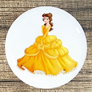 May include: A white round button with a cartoon image of Belle from Disney's Beauty and the Beast. Belle is wearing a yellow ball gown with a white lace trim and a yellow ribbon around her waist. She has brown hair and green eyes.