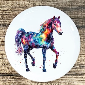 May include: A white, round decorative plate featuring a watercolor painting of a horse. The horse's body is a vibrant galaxy of colors, including blue, purple, orange, and yellow. The horse is depicted with its front leg raised.