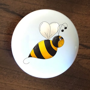 May include: A white ceramic knob with a cartoon bee design. The bee is yellow and black with a white heart-shaped wing. The bee has a smiling face and is flying.