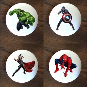 May include: Four white ceramic round knobs with images of popular superheroes. The top left knob features the Hulk, the top right knob features Captain America, the bottom left knob features Thor, and the bottom right knob features Spiderman.