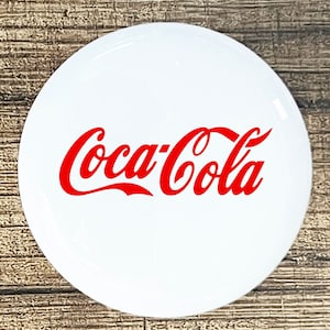 May include: A white round button with the red Coca-Cola logo.
