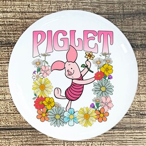 May include: A white round button with a pink gradient background featuring the word "Piglet" in a retro font. A cartoon image of Piglet from Winnie the Pooh is holding a flower and standing in a circle of colorful flowers.