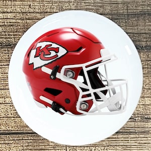 May include: A red and white Kansas City Chiefs football helmet with a white facemask. The helmet features the Chiefs logo on the side, set on a white plate. The background is a wooden surface.