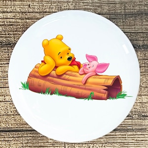 May include: Winnie the Pooh and Piglet are relaxing on a brown log in a green grassy area. Pooh is yellow and has a red shirt. Piglet is pink and has a pink nose.