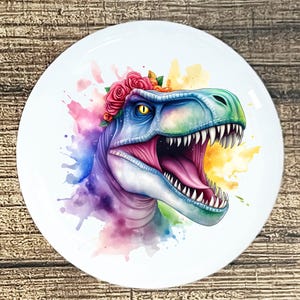 May include: A white ceramic plate featuring a watercolor illustration of a dinosaur head. The dinosaur is blue, green, and purple, with a pink mouth and a crown of red roses. The background has colorful watercolor splashes.