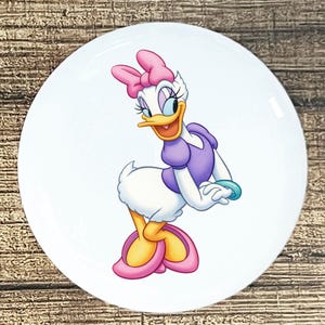 May include: A white ceramic plate featuring a cartoon image of Daisy Duck. Daisy is depicted with a pink bow, purple dress, and pink shoes. The plate has a glossy finish and is set against a wooden background.