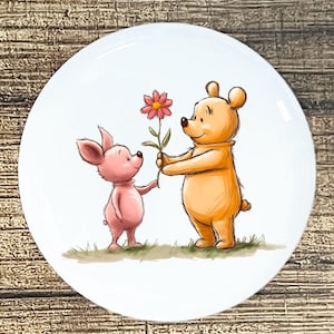 May include: A white ceramic plate featuring a charming illustration of Winnie the Pooh and Piglet. Pooh, in orange, offers a pink flower to Piglet, who is pink. The characters stand on green grass, creating a heartwarming scene.