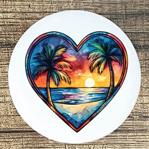 May include: A white, round ceramic plate featuring a heart-shaped design with a beach scene. The artwork depicts two palm trees, a sunset, and ocean waves. The color palette includes blue, orange, yellow, and green.