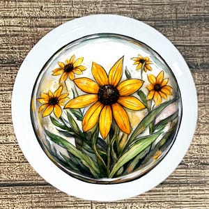 May include: A white, round decorative plate featuring a watercolor painting of yellow and brown Black-Eyed Susan flowers. The flowers are surrounded by green leaves and are enclosed in a glass-like bubble.