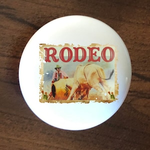 May include: A white ceramic knob with a vintage-style image of a rodeo scene. The image features a cowboy riding a bull with the word "RODEO" in red letters at the top.
