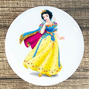 May include: A white ceramic plate with a colorful illustration of Snow White, a Disney princess, wearing a yellow dress with a blue bodice, a red cape, and a gold crown. She is smiling and holding her hand to her chin.