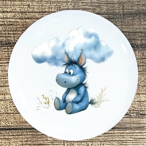 May include: A white ceramic plate featuring a whimsical illustration of a blue donkey sitting under a blue cloud. The donkey has a friendly expression and is surrounded by small patches of grass. The plate has a glossy finish.