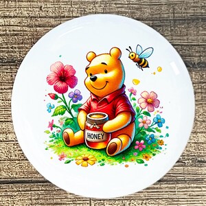 May include: A white ceramic plate featuring a smiling yellow bear in a red shirt, holding a honey jar. The plate is decorated with colorful flowers, green grass, and a flying bee. The word "HONEY" is visible on the jar.