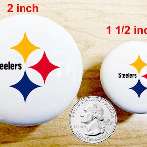 May include: Two white, round buttons with the Pittsburgh Steelers logo, featuring a gold, red, and blue starburst design and the word "Steelers". The larger button is 2 inches in diameter, and the smaller is 1 1/2 inches. A quarter is included for size comparison.