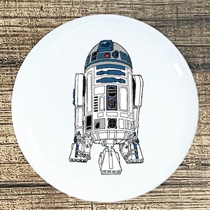 May include: A white, blue, and gray cartoon illustration of R2-D2, a robot from the Star Wars franchise. The robot is standing on a white background.