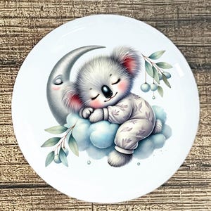 May include: A white ceramic plate featuring a sleeping koala bear on a cloud, resting against a crescent moon. The koala wears a patterned onesie. The design includes green leaves and blue berries, creating a whimsical, dreamy theme.