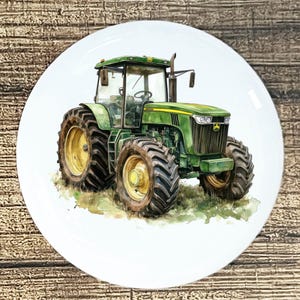 May include: A white ceramic plate featuring a watercolor painting of a green tractor with yellow wheels. The tractor is depicted on a grassy field. The plate has a glossy finish and is set against a wooden background.
