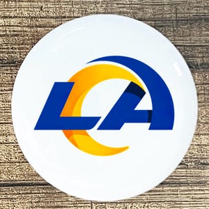 May include: A white, circular coaster featuring the Los Angeles Rams logo. The logo includes the letters "LA" in blue, with a yellow and orange curved design. The coaster is glossy and appears to be made of ceramic or a similar material.