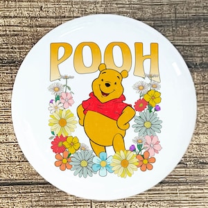 May include: A white round button with a yellow gradient "POOH" text at the top. Winnie the Pooh, a cartoon bear, is standing in the center of a circle of colorful flowers.