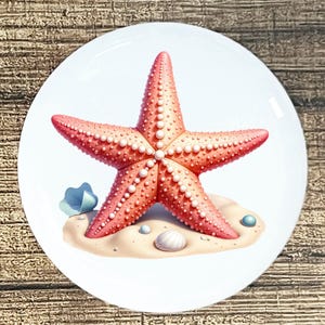 May include: A decorative plate featuring a coral-colored starfish with white pearl-like accents. The starfish rests on a sandy beach scene with seashells and a blue clam shell. The plate has a white background.