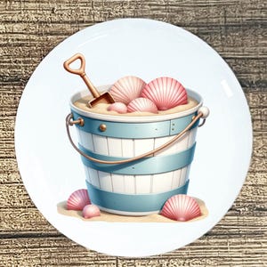 May include: A white ceramic plate featuring a beach scene. A blue and white striped bucket is filled with sand, pink seashells, and a small shovel. Several pink seashells are scattered around the bucket on a sandy surface.