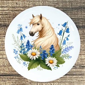 May include: White ceramic plate featuring a watercolor illustration of a horse with a flowing mane, surrounded by bluebell flowers and daisies. The horse is light tan, and the flowers are blue and white.