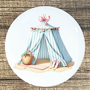 May include: A decorative plate featuring a beach scene. A blue and white striped tent is set on a sandy beach, with a pink mat, a woven basket, and pink flip-flops. The tent is adorned with pink bows.