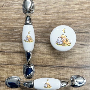 May include: Three ceramic cabinet knobs with a Winnie the Pooh and Piglet design. The knobs are white with a silver metal base. The design features Winnie the Pooh and Piglet hugging under a crescent moon.