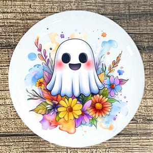 Puede incluir: Un plato de cerámica blanco con un diseño de fantasma de dibujos animados sonriente, con mejillas rosadas, rodeado de coloridas flores y follaje. El diseño incluye elementos de acuarela amarillos, naranjas, morados y azules.