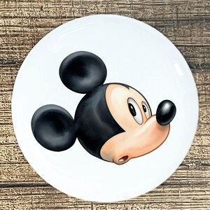 May include: A white ceramic plate featuring a side profile of Mickey Mouse. The cartoon character has black ears, a black nose, and a peach-colored face. The plate is round and has a glossy finish.