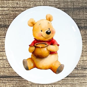May include: A white ceramic plate featuring a watercolor illustration of Winnie the Pooh. The bear is depicted in a seated position, wearing a red shirt and holding a honey pot. The plate has a glossy finish and a round shape.