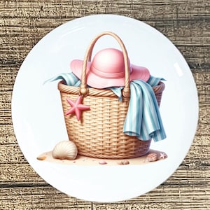 May include: A decorative plate featuring a beach scene. A woven picnic basket holds a pink sun hat, a blue and white striped towel, and a pink starfish charm. A seashell and sand complete the beach theme.