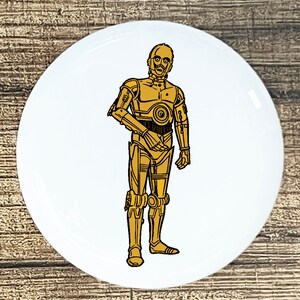 May include: A white button with a gold illustration of C-3PO, a character from Star Wars, standing with his arms at his sides.