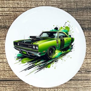 May include: A white ceramic plate featuring a green and black illustration of a classic Dodge muscle car. The car has black racing stripes and a large engine. Green and black paint splatters create a sense of motion.