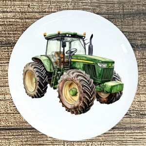 May include: A white ceramic plate featuring a watercolor illustration of a green John Deere tractor. The tractor has large tires, a cab, and the John Deere logo. The plate is round and set against a wooden background.